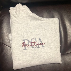 Ash gray 1/4 quarter zip sweatshirt with custom embroidery featuring PCA in steel gray in all caps with name Brittnee in cursive font in maroon thread.