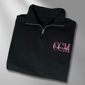May include: A black quarter-zip pullover with a silver zipper. The pullover features the embroidered text "CCM Deborah" in pink script on the front. The garment is made of a soft material and is folded neatly.