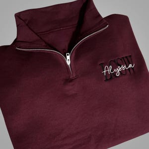 May include: Burgundy quarter-zip sweatshirt with a white embroidered name "Alyssa" on the right side of the chest.