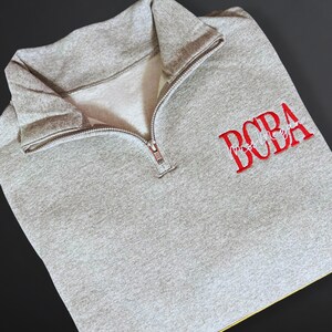 May include: A heather gray quarter-zip sweatshirt with the word "BOBA" embroidered in red, with "Mostly Boba" in a script font underneath. The sweatshirt has a silver zipper and a folded collar.