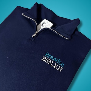 May include: Navy blue quarter-zip pullover with a silver zipper. The embroidered text "Branden BSN, RN" is in white and teal. A white tag with the text "Jane & Bradley" is tucked into the collar.