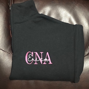 Black 1/4 quarter zip sweatshirt with custom embroidery featuring CNA in bubble gum pink in all caps with name Erica in cursive font in white thread.