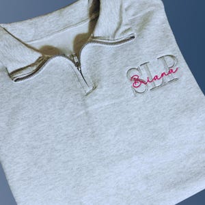 Ash color quarter zip with left chest embroidery featuring SLP credentials in capital serif font in light gray thread.  Name, Briana, in script font is embroidered through the center in fuschia thread.