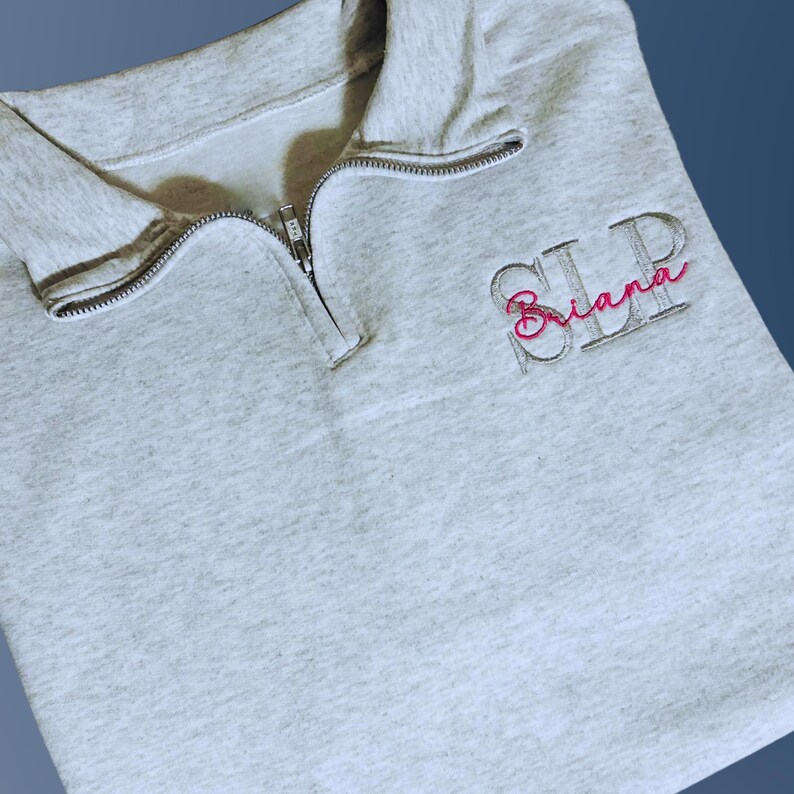 Ash color quarter zip with left chest embroidery featuring SLP credentials in capital serif font in light gray thread.  Name, Briana, in script font is embroidered through the center in fuschia thread.