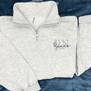 Ash gray 1/4 quarter zip sweatshirt with custom embroidery featuring RN in light gray in all caps with name Renee in cursive font in smokey blue thread.