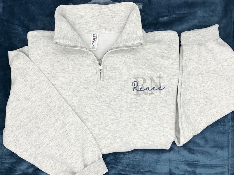 Ash gray 1/4 quarter zip sweatshirt with custom embroidery featuring RN in light gray in all caps with name Renee in cursive font in smokey blue thread.