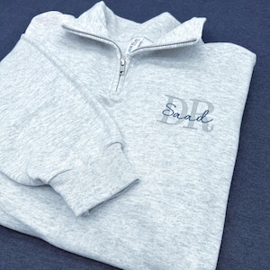 May include: A light gray sweatshirt with a quarter-zip collar. The sweatshirt features the embroidered name "Saad" in a dark blue script, with the initials "DR" above the name. The fabric has a heathered appearance.
