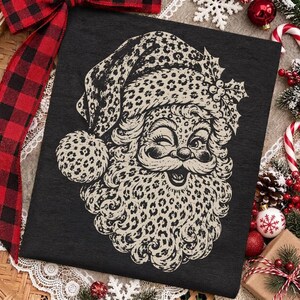 Op de afbeelding: Een zwart t-shirt met een grafische print van een knipogend gezicht van de Kerstman. Het ontwerp van de Kerstman is gevuld met een beige en zwart luipaardpatroon, inclusief de muts, baard en snor, met hulstbladeren en bessen op de muts.