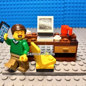 Op de afbeelding: Een Lego minifiguur met een groene trui en een bruine broek zit aan een Lego bureau met een computer, een kopje en een stoel. De minifiguur houdt een mobiele telefoon vast. Het bureau is bruin met een lade en een plank. De stoel is geel.