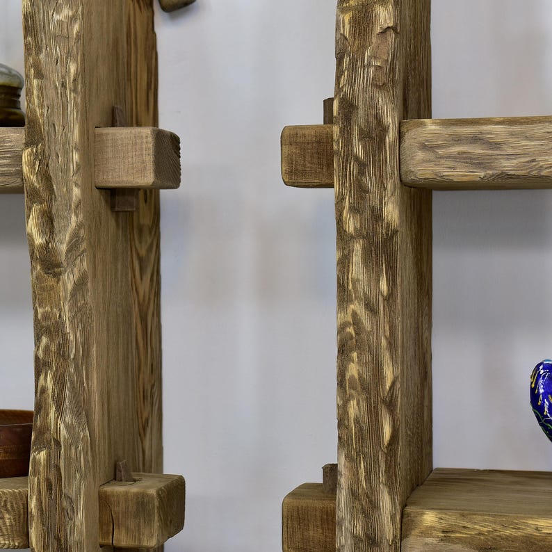 May include: A rustic wooden shelf unit with a natural, weathered finish. The shelves are supported by thick, vertical posts and smaller wooden blocks. The wood grain is visible, adding to the natural aesthetic. The design is simple and sturdy.