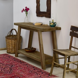 May include: A rustic wooden console table with a lower shelf, holding a small wooden bowl. A wicker basket and a wooden chair are nearby. The table is topped with a small lamp, books, and decorative items. A red patterned rug is in the foreground.