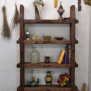 May include: A rustic, dark brown wooden bookshelf with four shelves, displaying various decorative items. These include vases, books, a lamp, and small cups. Dried flowers and other ornaments are also visible, creating a warm, inviting atmosphere.
