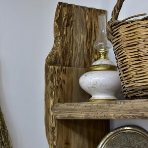 May include: A rustic wooden shelf with a weathered appearance, holding a white and gold oil lamp, a woven basket, and a decorative silver tray. The shelf is mounted against a white wall, creating a contrast with the warm tones of the wood.