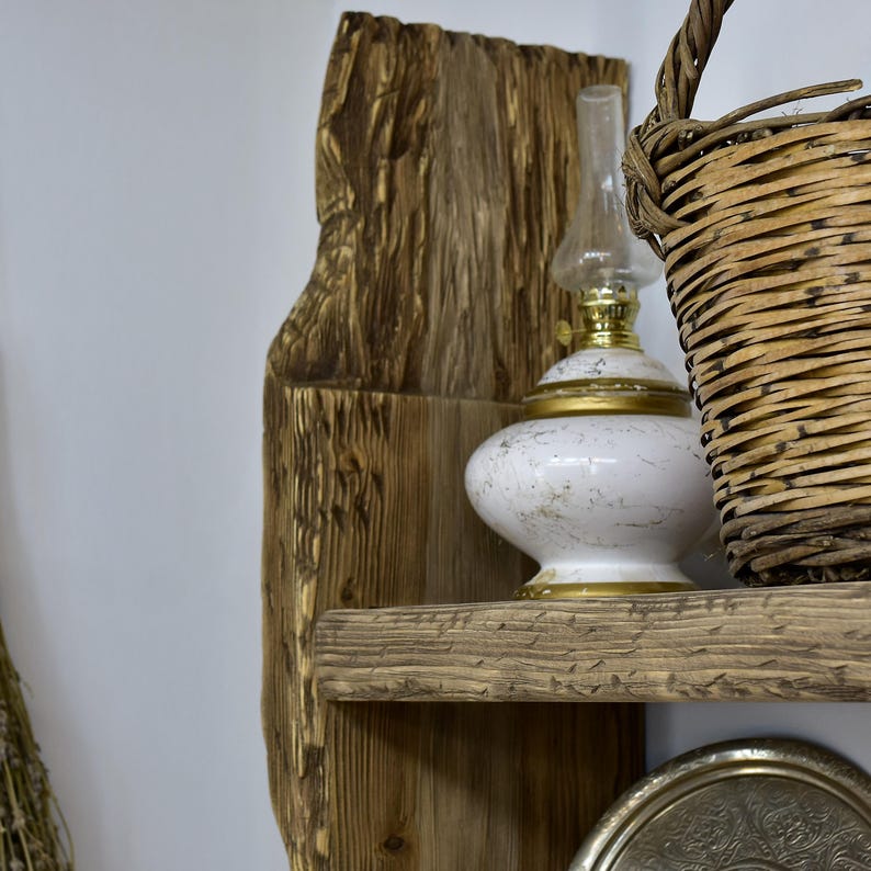 May include: A rustic wooden shelf with a weathered appearance, holding a white and gold oil lamp, a woven basket, and a decorative silver tray. The shelf is mounted against a white wall, creating a contrast with the warm tones of the wood.