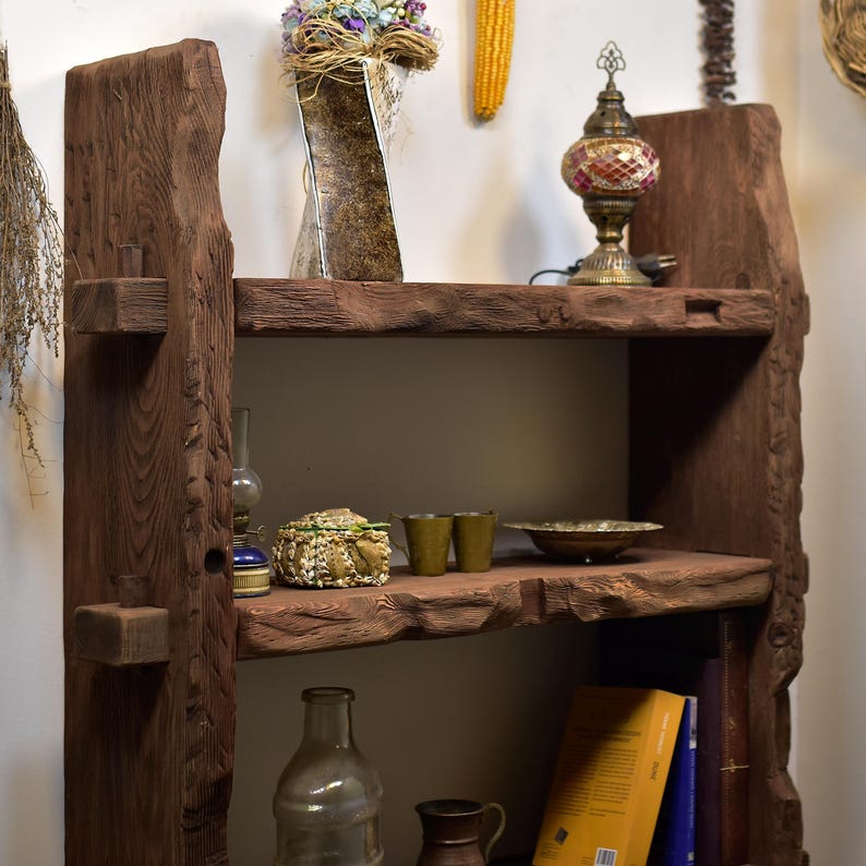 May include: Rustic, dark brown wooden bookshelf with two shelves. The shelves hold various decorative items, including a mosaic lamp, a vase of dried flowers, and books. The wood has a natural, unfinished look.
