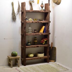 May include: A rustic, dark brown wooden bookshelf with five shelves. The shelves display various decorative items, including vases, books, and a small stool with a green gourd. Dried herbs and other decor hang on the wall.