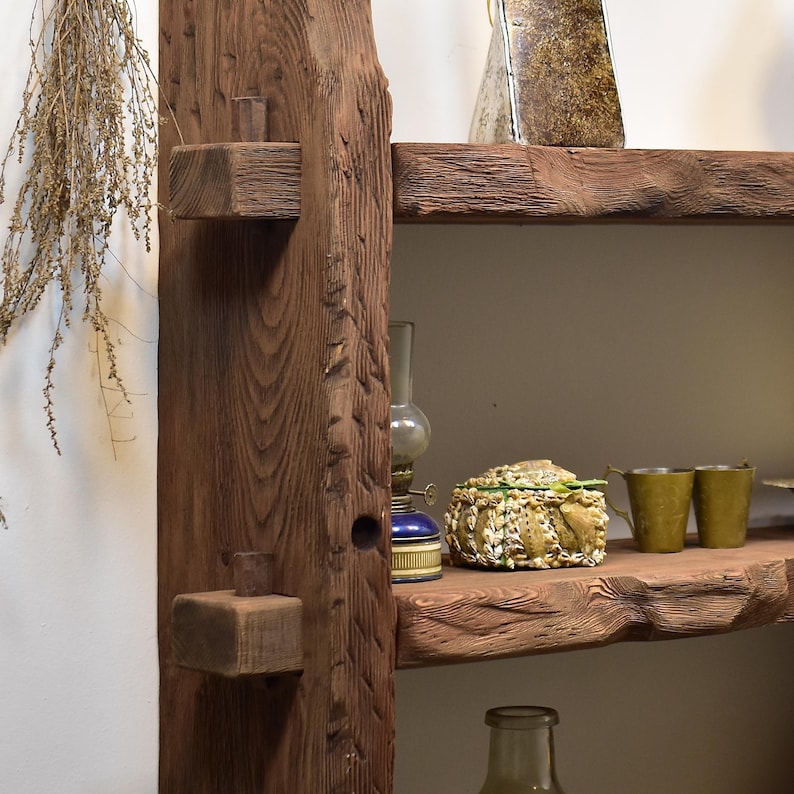 May include: A rustic wooden shelf unit with two shelves and a vertical support. The shelves are made of rough-hewn wood, and the unit features a natural, dark brown finish. Decorative items, including a lamp and small cups, are displayed on the shelves.