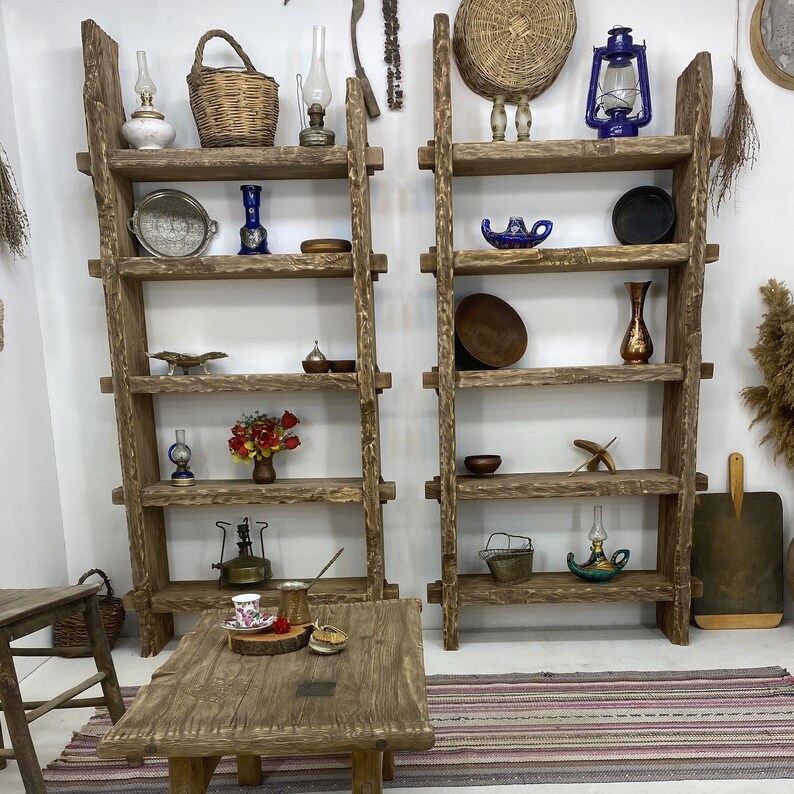 May include: Two rustic wooden ladder shelves with various decorative items. The shelves are filled with vintage lanterns, baskets, vases, and small bowls. A wooden table with a coffee set sits in front of the shelves, creating a cozy, vintage aesthetic.