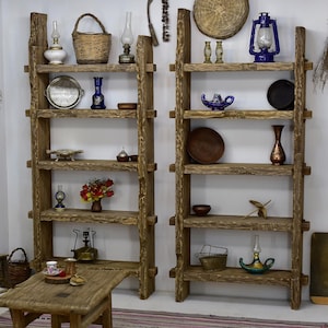 May include: Two rustic wooden ladder shelves filled with decorative items. The shelves are made of rough-hewn wood and feature various objects, including baskets, vases, lamps, and small dishes. The items are arranged on the shelves against a white wall.