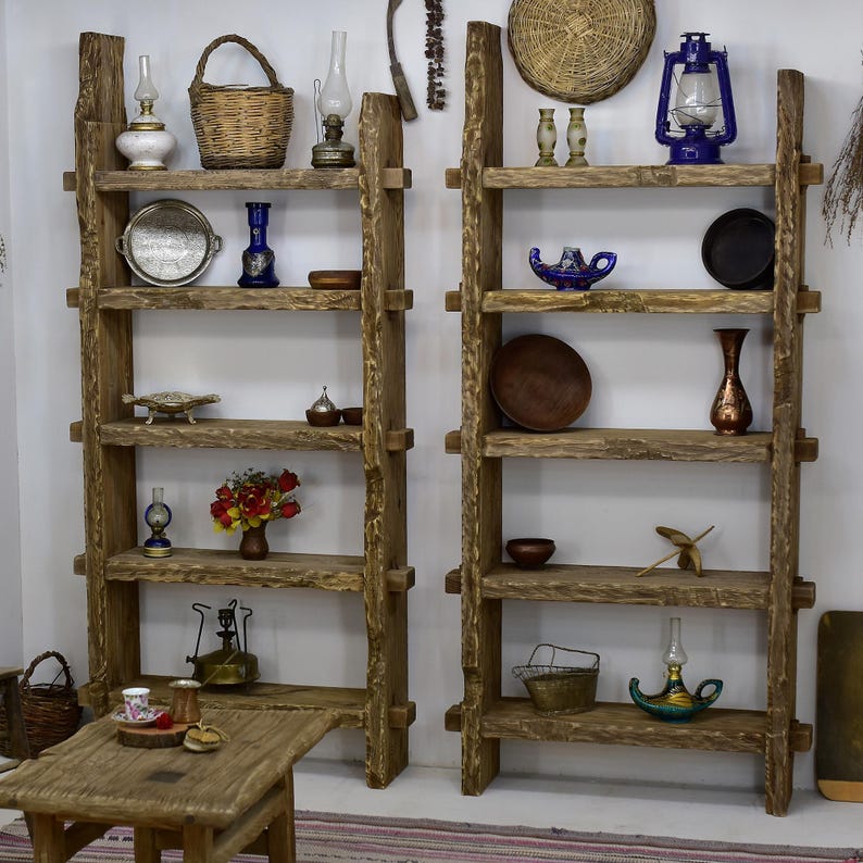 May include: Two rustic wooden ladder shelves filled with decorative items. The shelves are made of rough-hewn wood and feature various objects, including baskets, vases, lamps, and small dishes. The items are arranged on the shelves against a white wall.