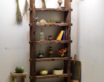 May include: A rustic, wooden bookshelf with multiple shelves, displaying various decorative items. The shelves hold a glass bottle, vases, books, and a small stool with a gourd. Dried herbs and other decor hang from the wall.