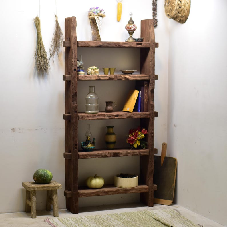 May include: A rustic, wooden bookshelf with multiple shelves, displaying various decorative items. The shelves hold a glass bottle, vases, books, and a small stool with a gourd. Dried herbs and other decor hang from the wall.