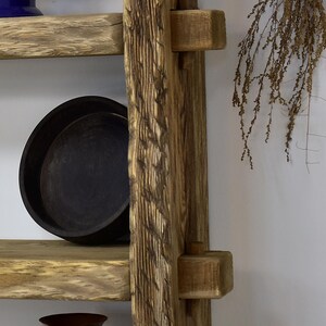May include: A rustic wooden shelf unit with a ladder-like design. The shelves are made of light-colored wood, and a black, round dish sits on one shelf. The unit has a natural, weathered appearance, with visible wood grain.