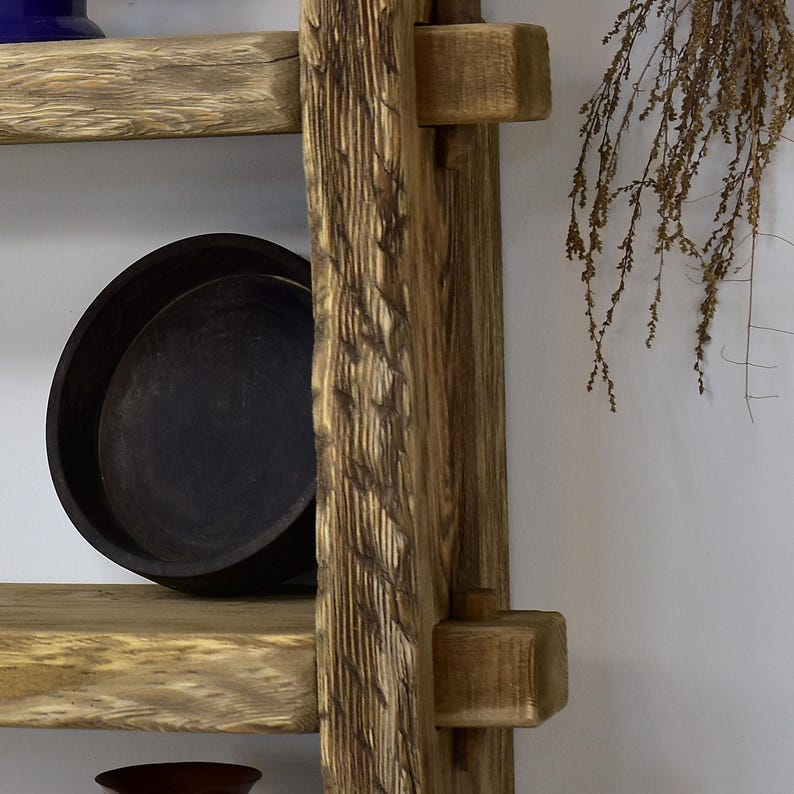 May include: A rustic wooden shelf unit with a ladder-like design. The shelves are made of light-colored wood, and a black, round dish sits on one shelf. The unit has a natural, weathered appearance, with visible wood grain.