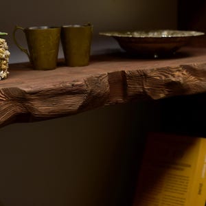 May include: A rustic wooden shelf displays two olive-green metal mugs with ornate handles and a decorative bowl. The shelf's edge has a natural, rough-hewn texture. The items are arranged on a dark, wooden surface.