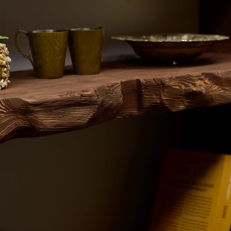 May include: A rustic wooden shelf displays two olive-green metal mugs with ornate handles and a decorative bowl. The shelf's edge has a natural, rough-hewn texture. The items are arranged on a dark, wooden surface.