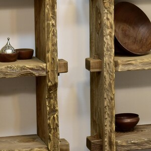May include: Rustic wooden shelves with a natural, unfinished appearance. The shelves are constructed from thick, textured planks and feature a warm, earthy tone. Decorative bowls and a small metal container with a lid are displayed on the shelves, adding a touch of elegance.