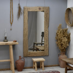 May include: A rustic wooden framed mirror with a rectangular shape and a natural wood finish. The mirror is hanging on a grey wall with a wooden stool and a clay jug in front of it.