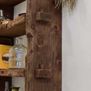 May include: A close-up of a rustic wooden shelf and support beam. The beam is a rich brown color with visible wood grain and two protruding wooden blocks. A glass bottle and a yellow box are visible on the shelf.