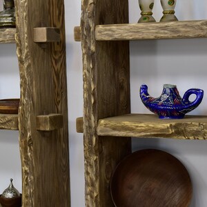 May include: Rustic wooden shelves with a natural edge design. A blue genie lamp with intricate patterns sits on one shelf. The shelves are made of light brown wood and are mounted on a white wall. A wooden bowl and decorative items are also visible.