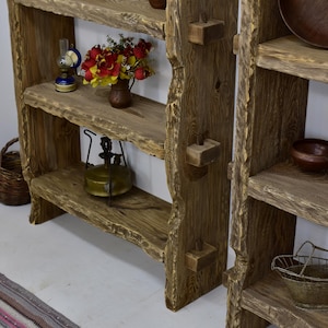 May include: Rustic wooden shelving unit with three shelves. The shelves are made of rough-hewn wood and are stained a warm brown. Decorative items include a small oil lamp, a vase of flowers, and a vintage lantern. The unit has a natural, organic aesthetic.