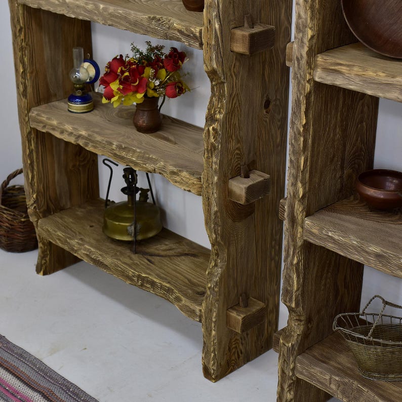 May include: Rustic wooden shelving unit with three shelves. The shelves are made of rough-hewn wood and are stained a warm brown. Decorative items include a small oil lamp, a vase of flowers, and a vintage lantern. The unit has a natural, organic aesthetic.