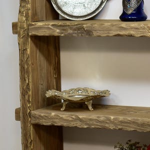 May include: A rustic wooden shelf unit with three shelves. The shelves hold decorative items, including a silver tray with intricate designs, a gold-colored decorative bowl, and a blue vase.