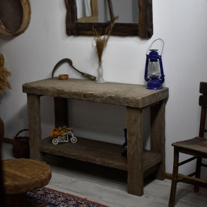 Pode incluir: Uma mesa consola de madeira rústica com uma única prateleira. A mesa tem um acabamento de madeira natural e é feita de madeira recuperada. A mesa é decorada com um pequeno carrinho de brinquedo, um vaso com flores secas e uma lamparina de óleo azul.