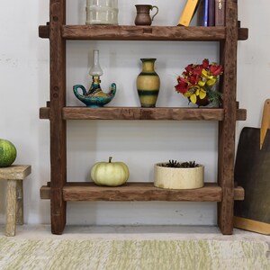 May include: A rustic, wooden shelf unit with three shelves, displaying various decorative items. The shelves hold a small pumpkin, a vase, a lamp, and a bouquet of flowers. The unit is made of dark brown wood.