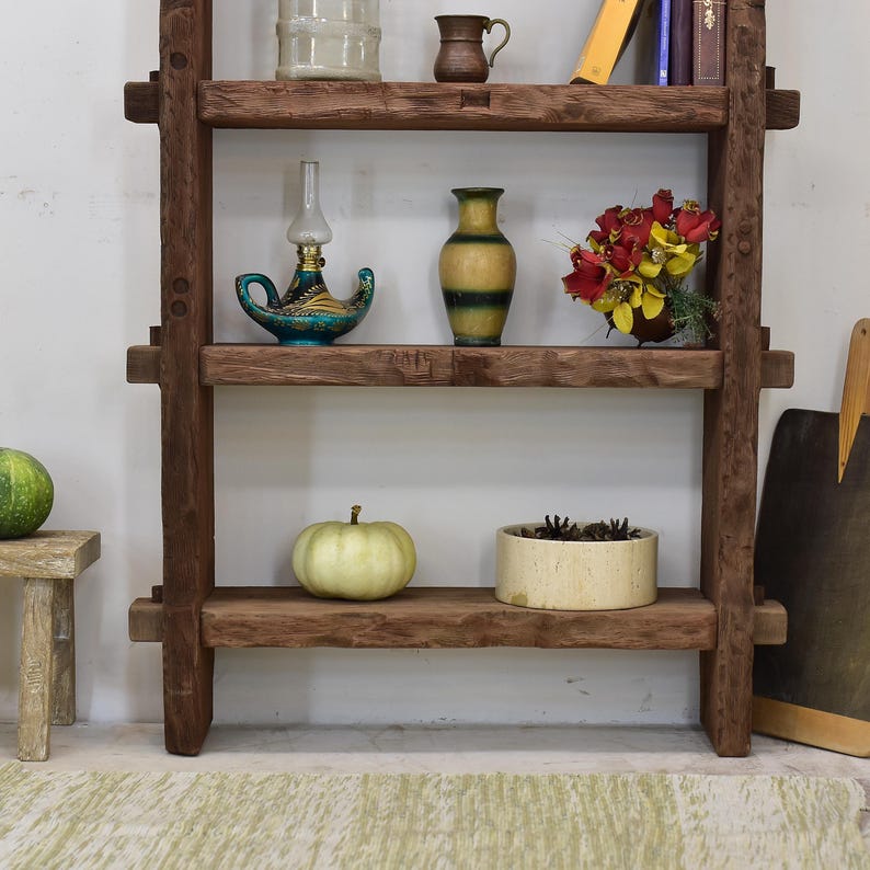 May include: A rustic, wooden shelf unit with three shelves, displaying various decorative items. The shelves hold a small pumpkin, a vase, a lamp, and a bouquet of flowers. The unit is made of dark brown wood.