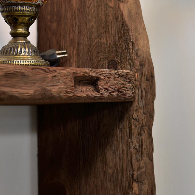 May include: A rustic, wooden shelf with a natural, textured finish. The shelf is part of a larger wooden structure, showcasing the raw, unfinished beauty of the wood. A decorative lamp with a brass base sits on the shelf, with a plug visible.