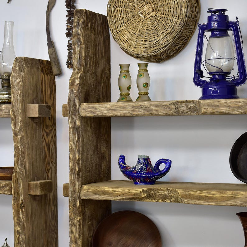 May include: A rustic wooden shelf unit with live edge shelves displays various decorative items. A blue oil lamp, a woven basket, and small glass vases are among the items. The shelves are supported by thick, unfinished wooden posts.