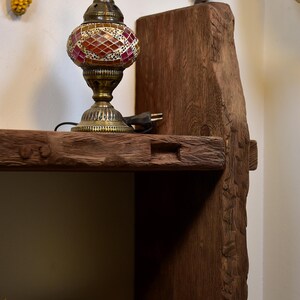 May include: A decorative table lamp with a mosaic glass shade in shades of red, orange, and gold sits on a rustic wooden shelf. The shelf is part of a larger wooden structure with visible grain and a natural, unfinished appearance. The lamp has a brass base.