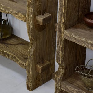May include: Close-up of a rustic wooden shelf unit with a natural, unfinished appearance. The shelves have a live edge design, showcasing the wood's natural grain and texture. Wooden pegs are used for structural support. The unit is a warm brown color.