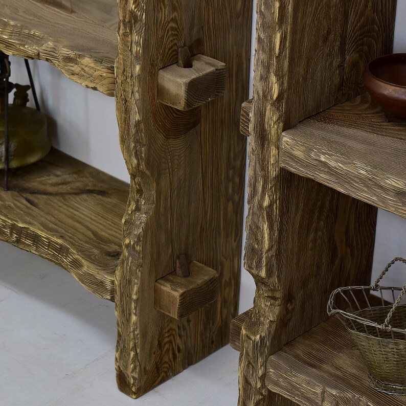 May include: Close-up of a rustic wooden shelf unit with a natural, unfinished appearance. The shelves have a live edge design, showcasing the wood's natural grain and texture. Wooden pegs are used for structural support. The unit is a warm brown color.