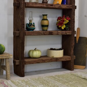 May include: A rustic, dark brown wooden shelf unit with three shelves. The shelves display a variety of decorative items, including a small oil lamp, a vase, a bowl of flowers, a pumpkin, and a container of pinecones. A small wooden stool is on the left.