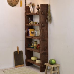 May include: A rustic, dark brown wooden bookshelf with four shelves, displaying various decorative items. The shelves hold books, vases, a glass bottle, and a small pumpkin. A wooden cutting board leans against the wall.