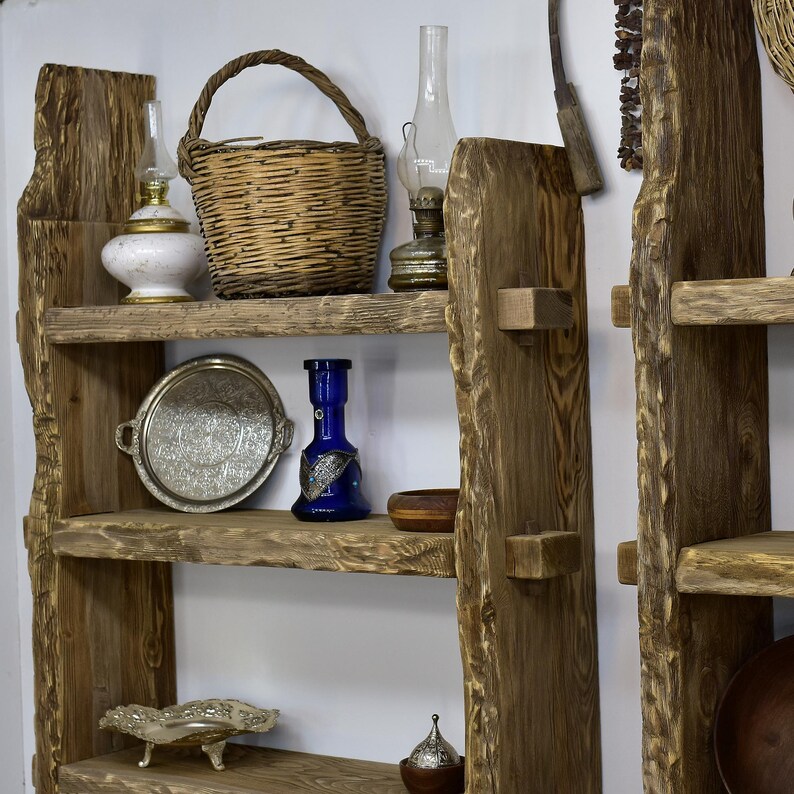 May include: A rustic wooden shelf unit with multiple shelves displaying various decorative items. Items include a woven basket, antique oil lamps, a silver tray, a blue glass bottle, and small wooden bowls. The shelves are made of rough-hewn wood.