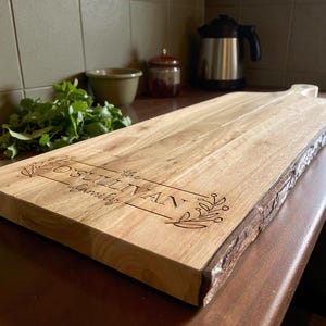 May include: A rectangular wooden cutting board with a natural edge and a handle. The board is engraved with the text "The O'Sullivan Family" and decorative floral elements. A lemon and herbs are in the background.