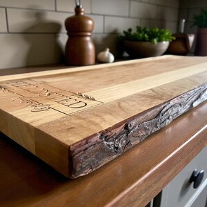 May include: A rectangular wooden cutting board with a live edge and the engraved text "FIELDS SHED". The board is light brown with a natural wood grain and a dark brown, textured edge. The board is on a wooden countertop.